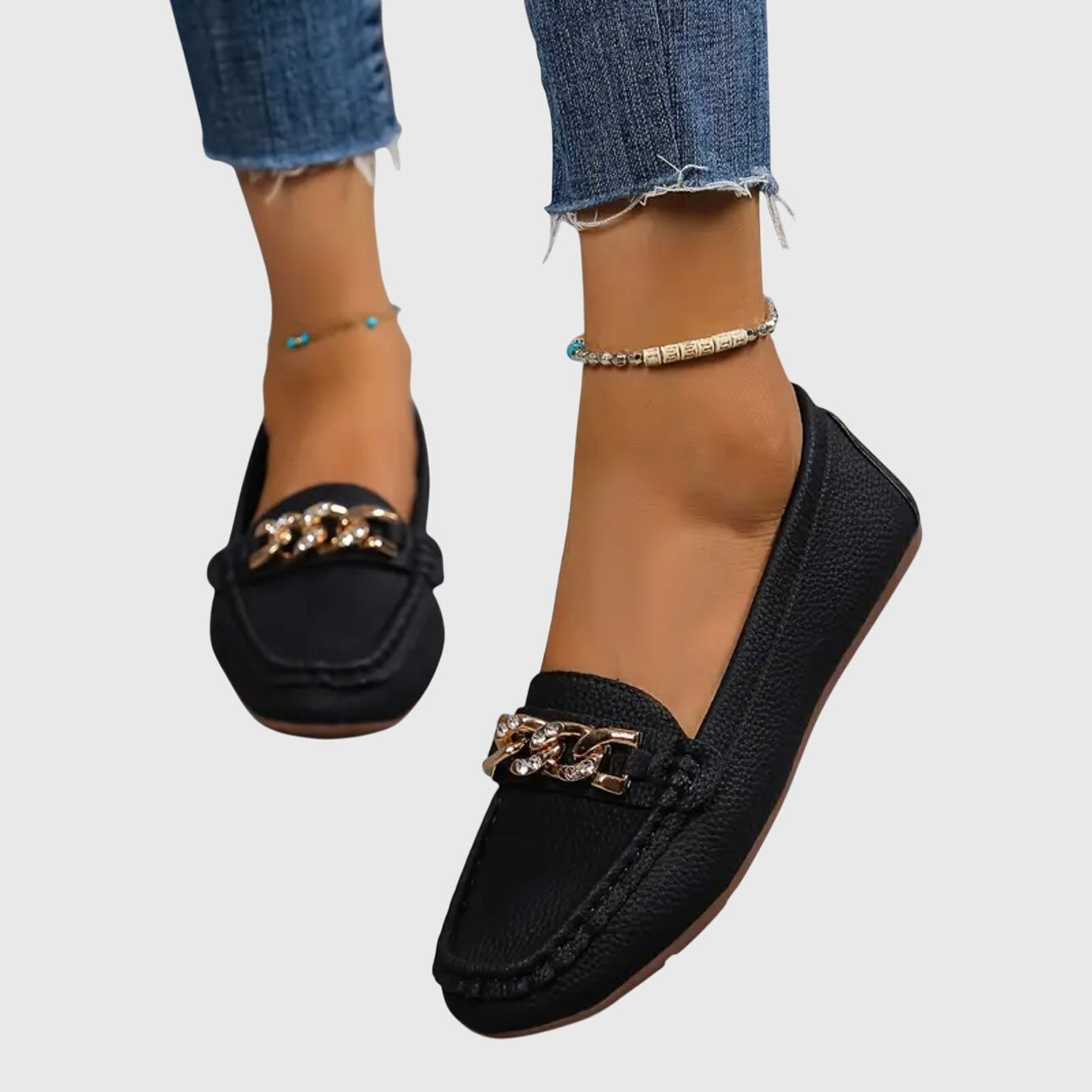 Black loafers with gold chain detail worn with blue jeans on a light gray background