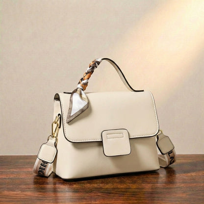 White leather crossbody bag with a structured top handle wrapped in a patterned scarf, placed on a wooden surface against a neutral background.