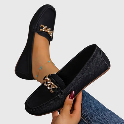 Black loafers with decorative straps held by a hand on a light gray background