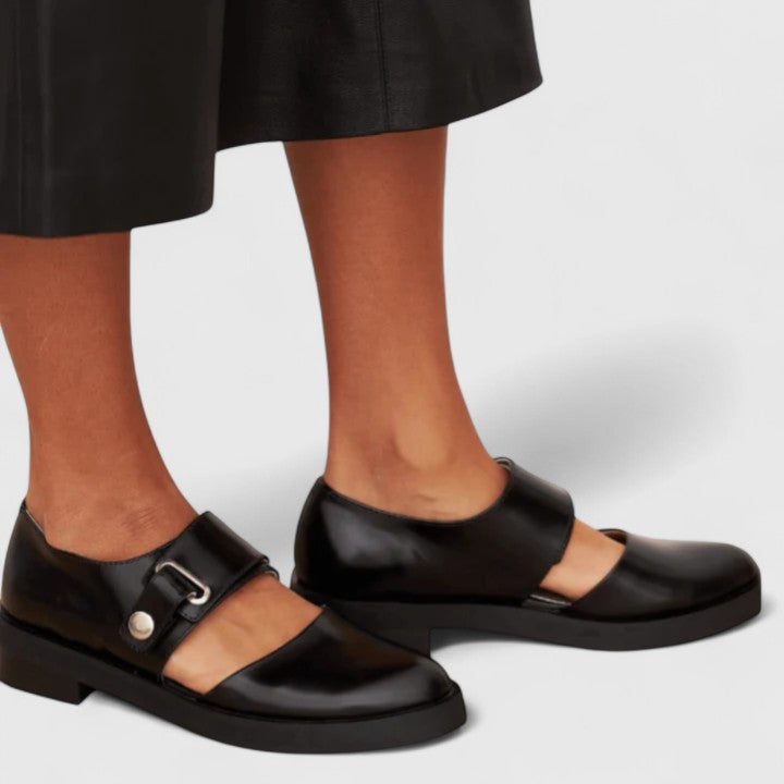 Black leather shoes with a buckle detail worn by a person on a light gray background