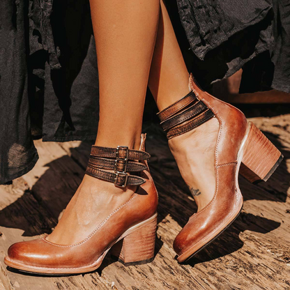 Brown leather ankle boots with strap details worn by a person on a wooden floor.