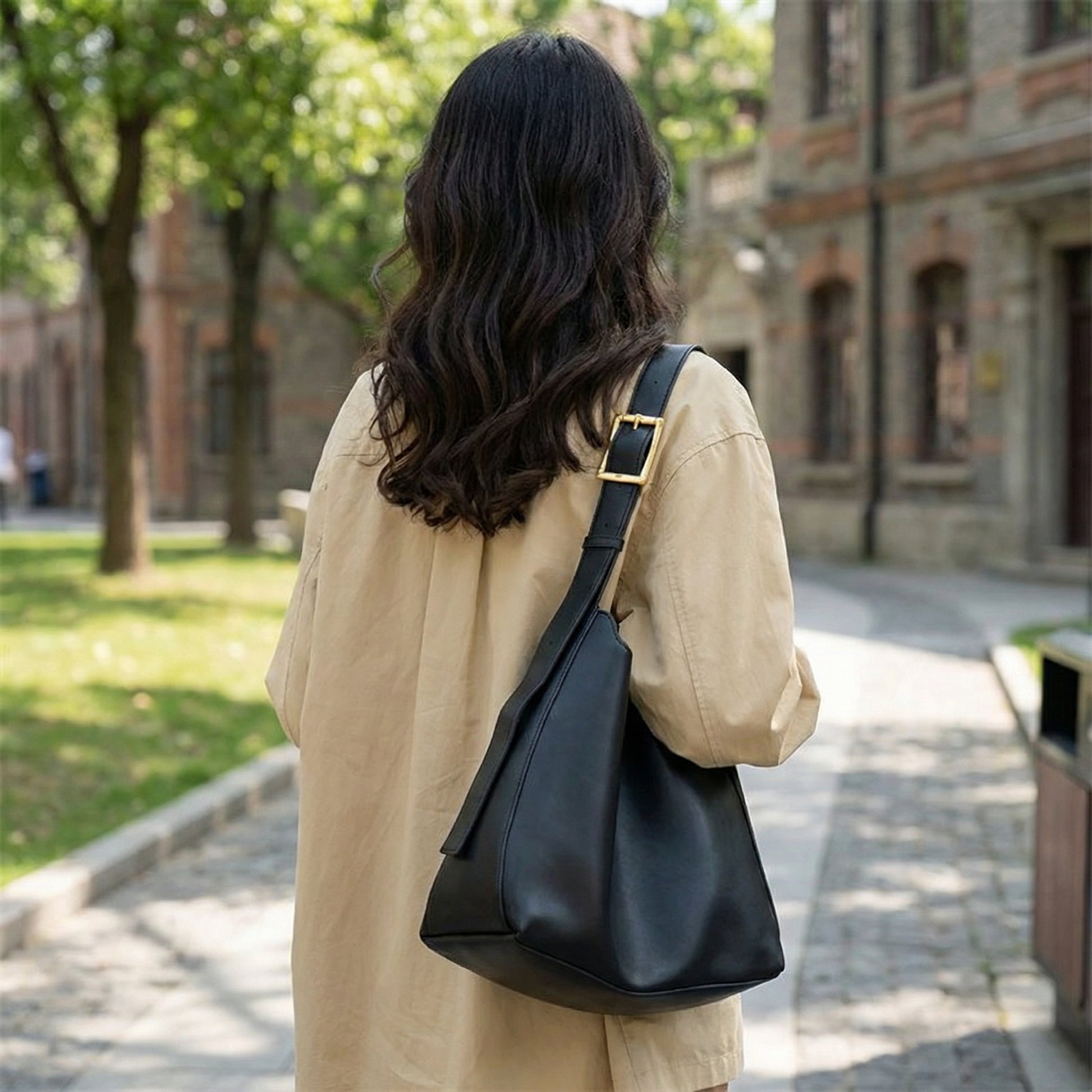 Woman wearing a black leather hobo shoulder bag with adjustable strap and gold buckle, soft minimalist purse styled for everyday city wear in an outdoor urban setting.