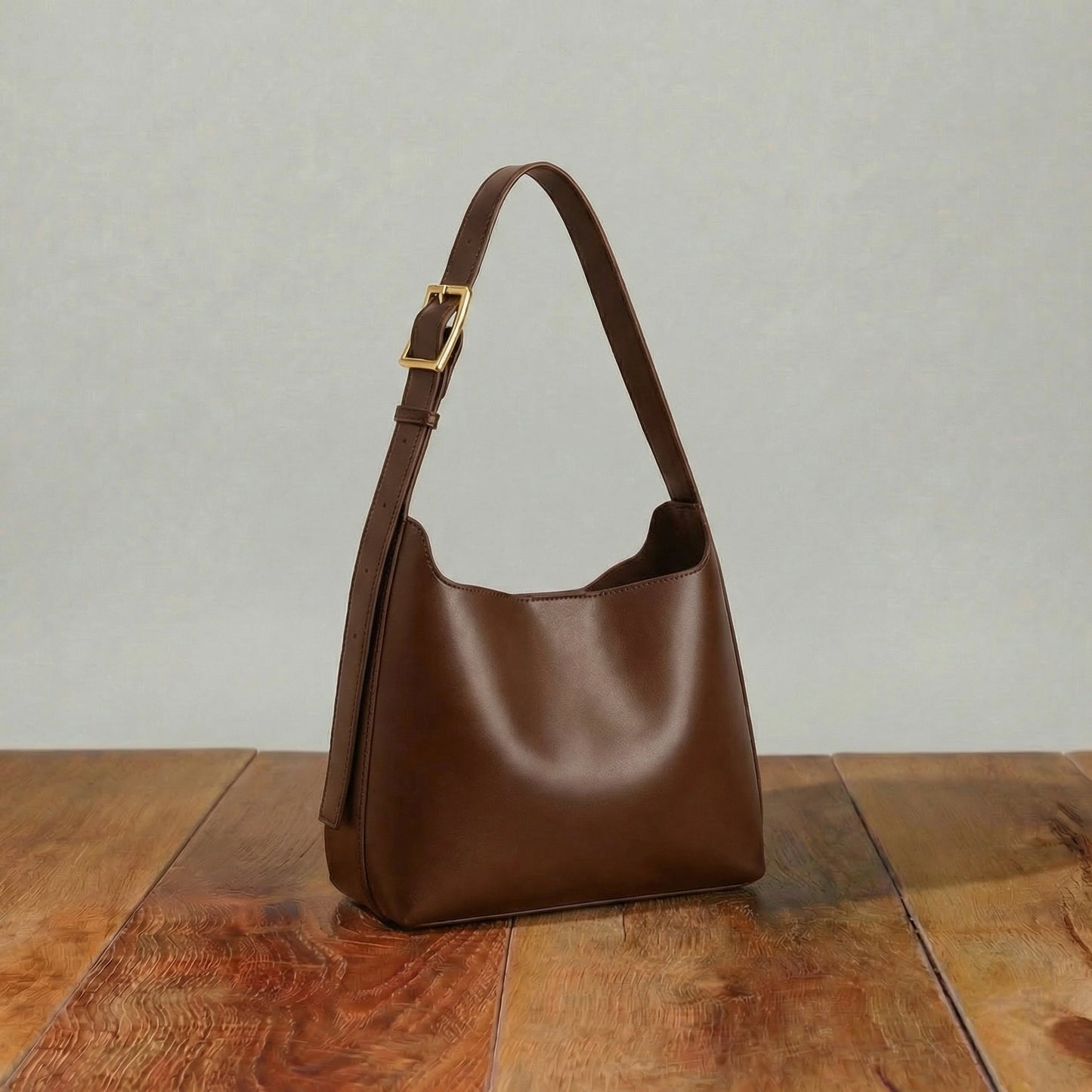 Brown leather hobo bag with soft curved shape and adjustable strap featuring gold buckle displayed on wooden table.
