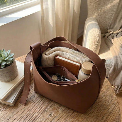 Open brown leather hobo bag with organized interior compartments, showing wallet, notebook, keys and essentials, minimalist shoulder purse styled on wooden table in cozy home setting.