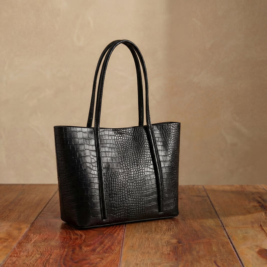 Black embossed leather tote bag with long shoulder handles displayed on wooden table.
