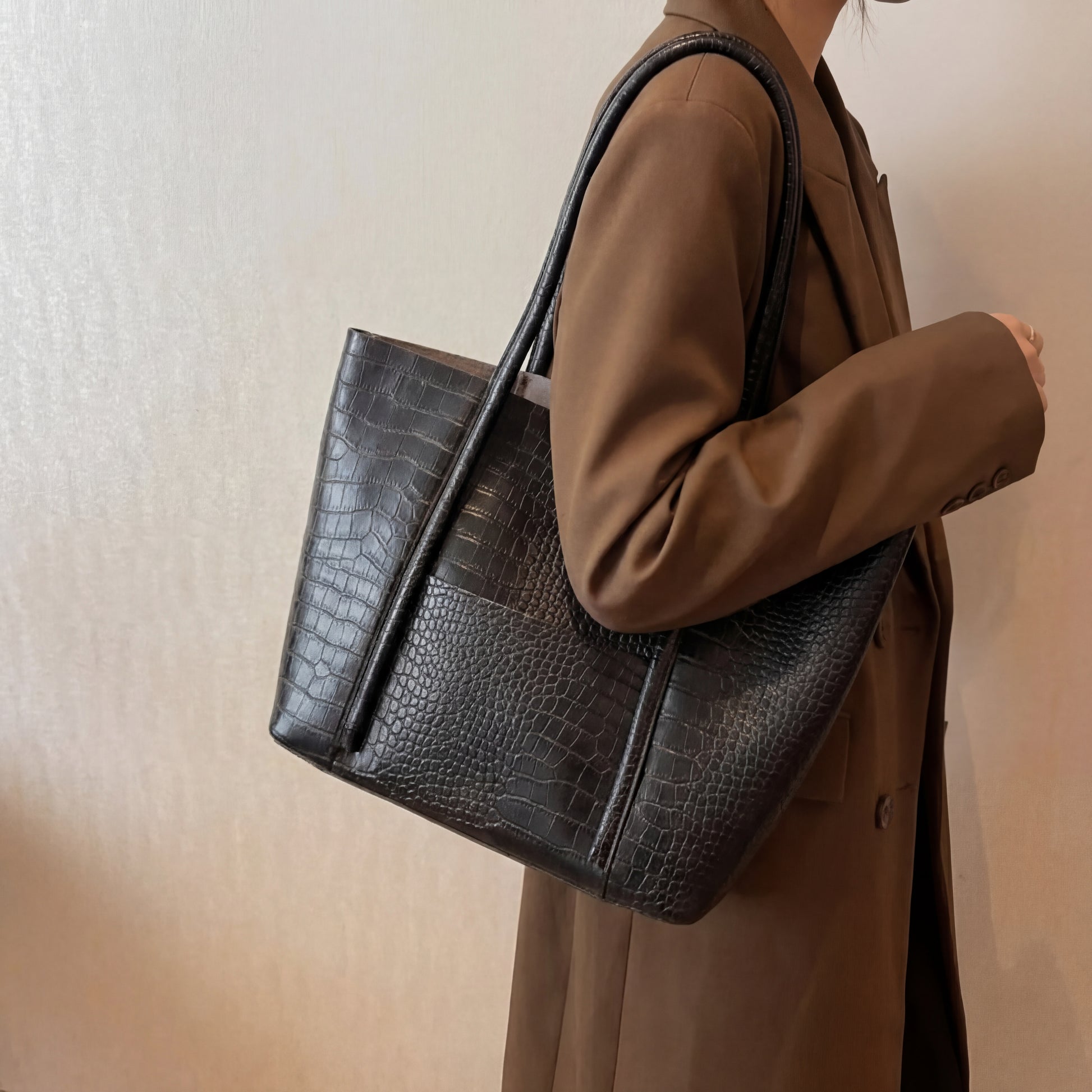 Woman carrying a black embossed leather tote bag over the shoulder, structured textured handbag styled with a brown blazer for a modern minimalist look.
