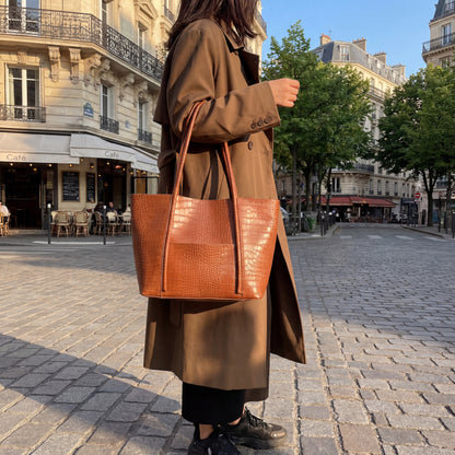 Person wearing a long brown coat and holding a brown tote leather bag on a city street.
