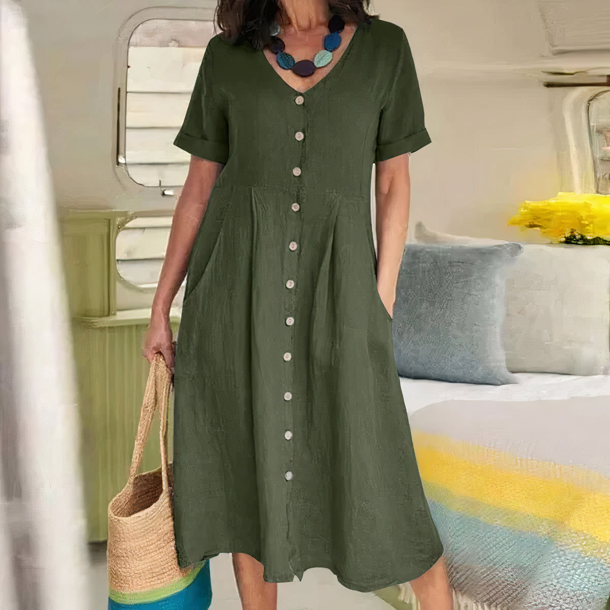 Woman wearing a green dress with buttons, holding a straw bag, in a room with a bed and pillows.