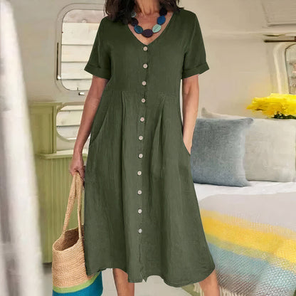 Woman wearing a green dress with buttons, holding a straw bag, in a room with a bed and pillows.