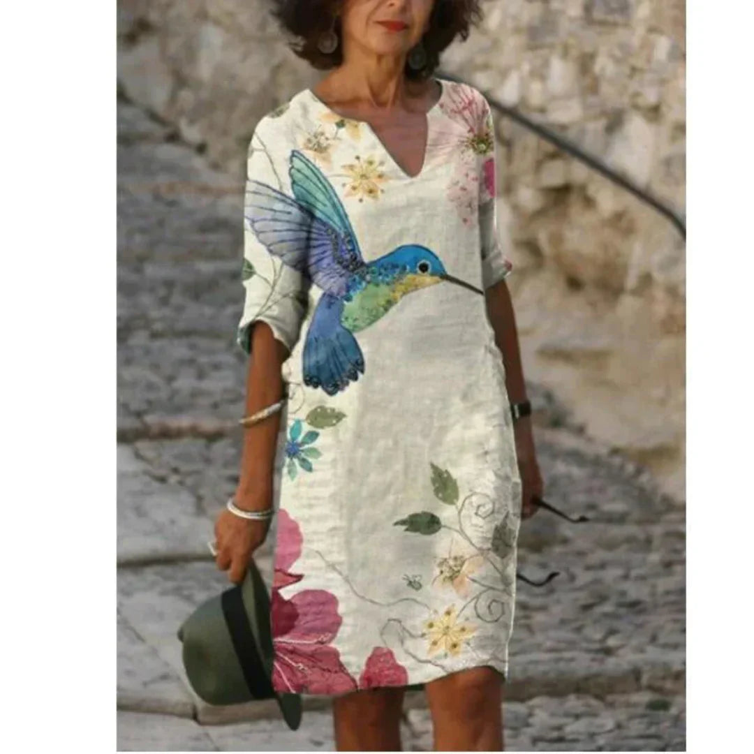 Woman wearing a floral dress with a colorful bird design, standing against a stone wall.