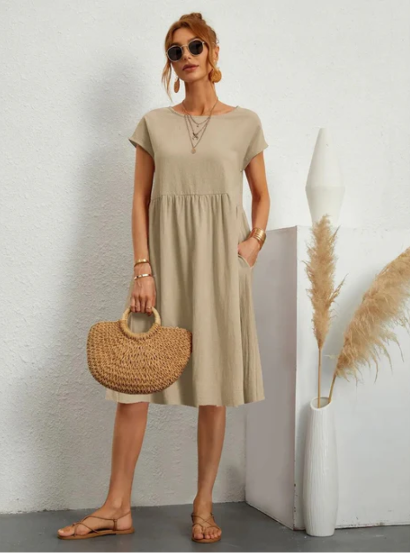 Woman wearing a beige dress holding a straw bag against a white wall.