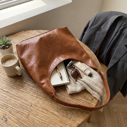 Light brown leather shoulder bag on a wooden table with a notebook, sunglasses, and a cup of coffee.
