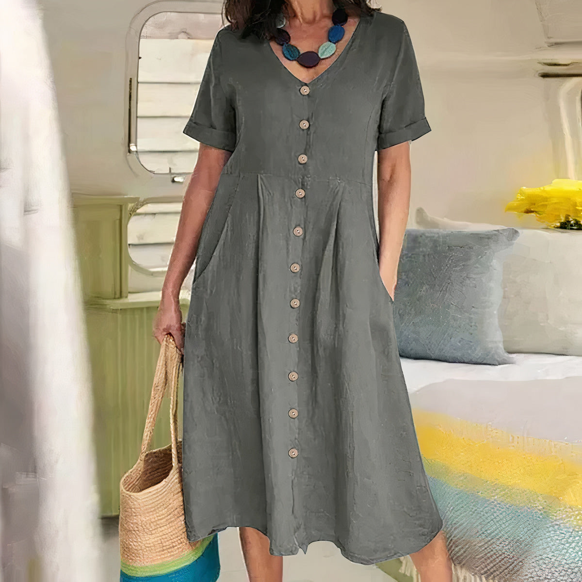 Woman wearing a gray dress with buttons, holding a straw bag, in a room with a bed and pillows.