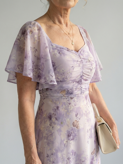 Woman wearing a light purple floral dress holding a beige clutch against a plain background