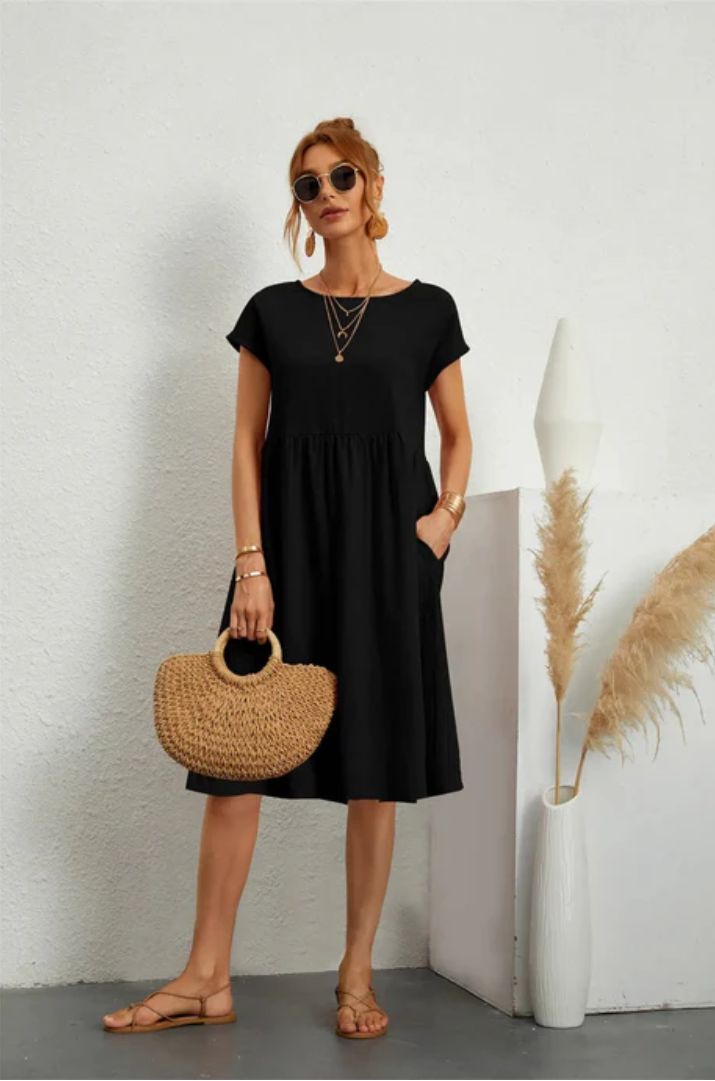 Woman wearing a black dress holding a straw bag against a white wall.