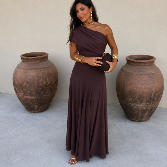 Woman in a brown one-shoulder dress standing between two large clay pots against a plain wall.