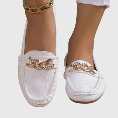 White loafers with gold chain details worn by a person on a light gray background