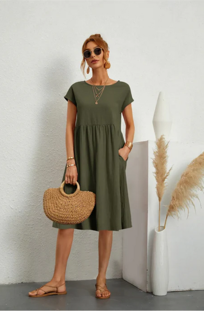 Woman wearing an olive green dress holding a straw bag against a white wall.