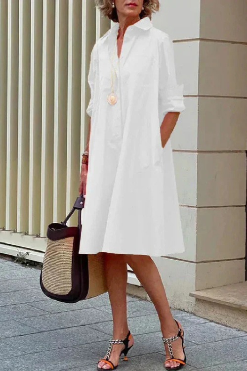 Woman wearing a white dress with a black and straw handbag, standing against a light-colored wall.