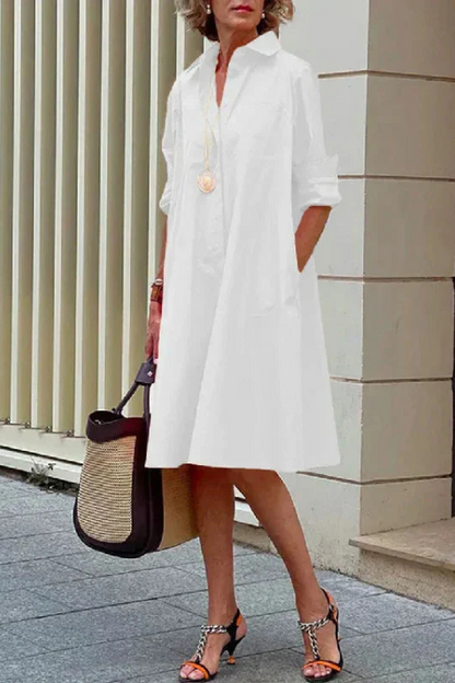 Woman wearing a white dress with a black and straw handbag, standing against a light-colored wall.