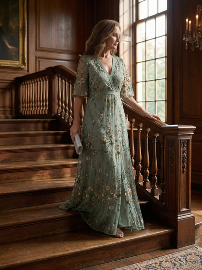 Woman in a green floral dress standing on wooden stairs in an elegant interior setting.