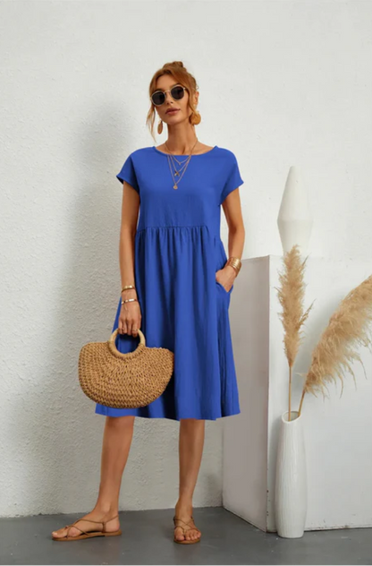 Woman wearing a blue dress holding a straw bag against a white wall.