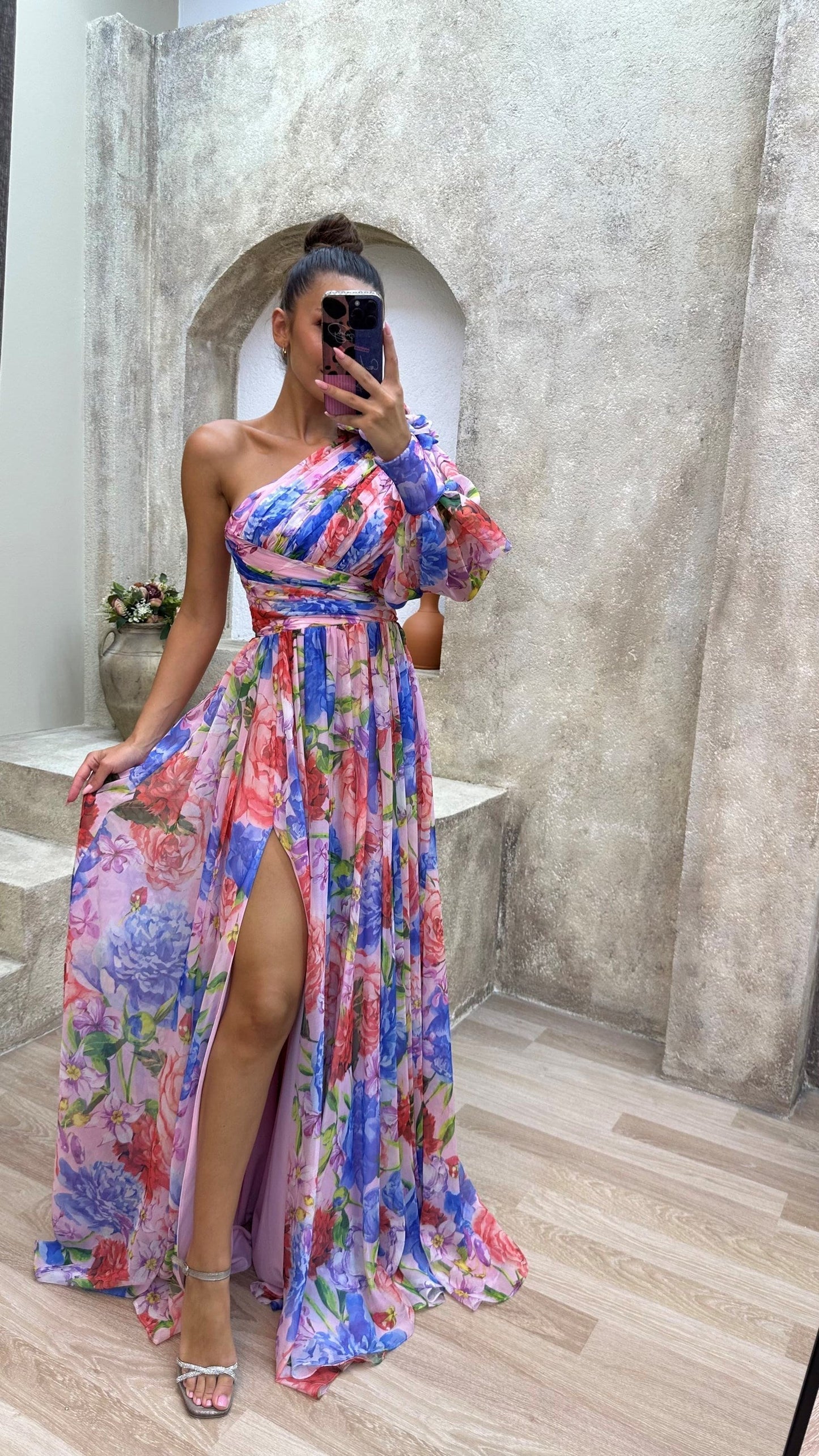 Woman wearing a colorful floral dress with a high slit, standing in front of a stone wall.