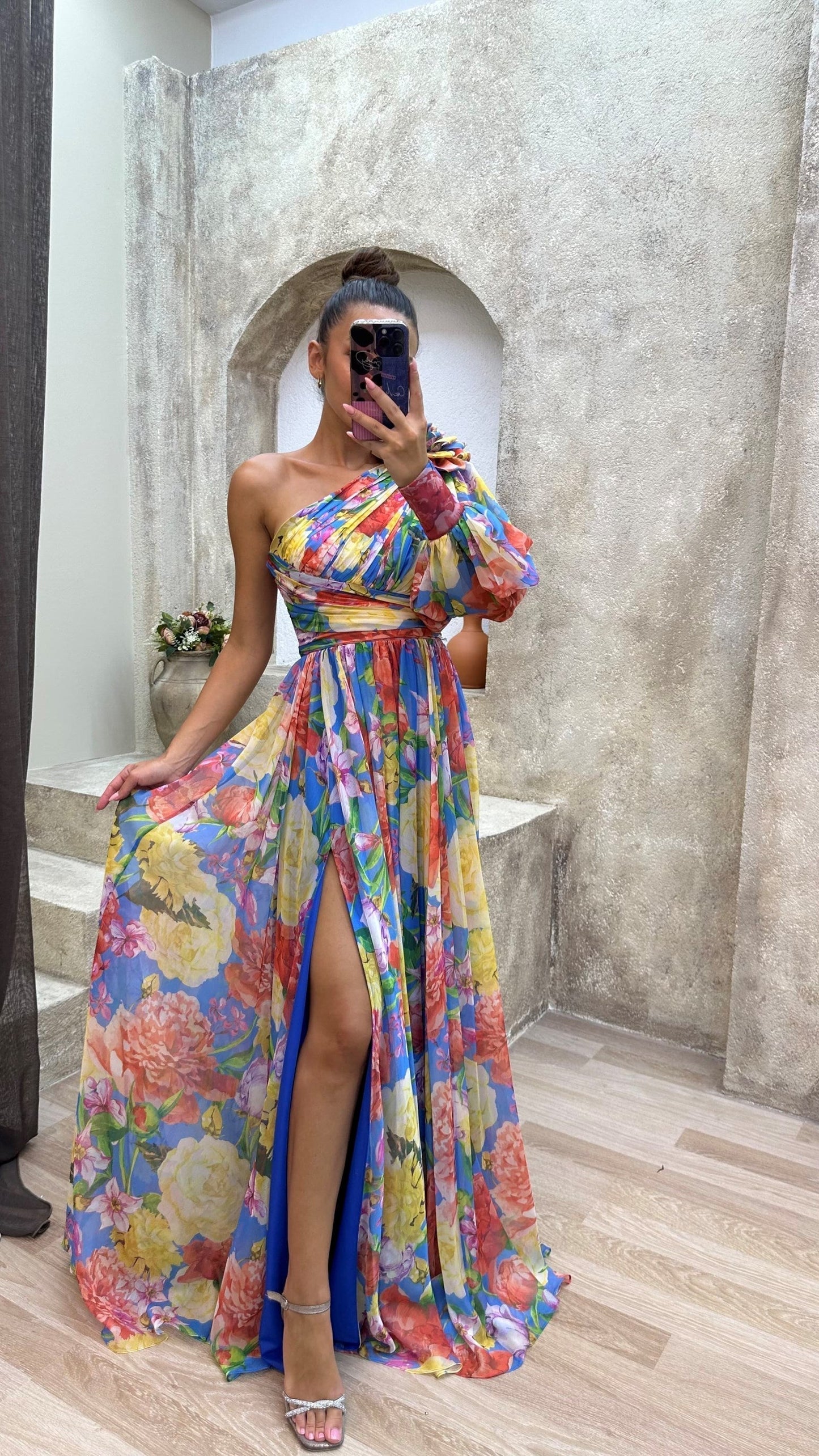 Person wearing a colorful floral dress with a high slit, standing in front of a stone wall.