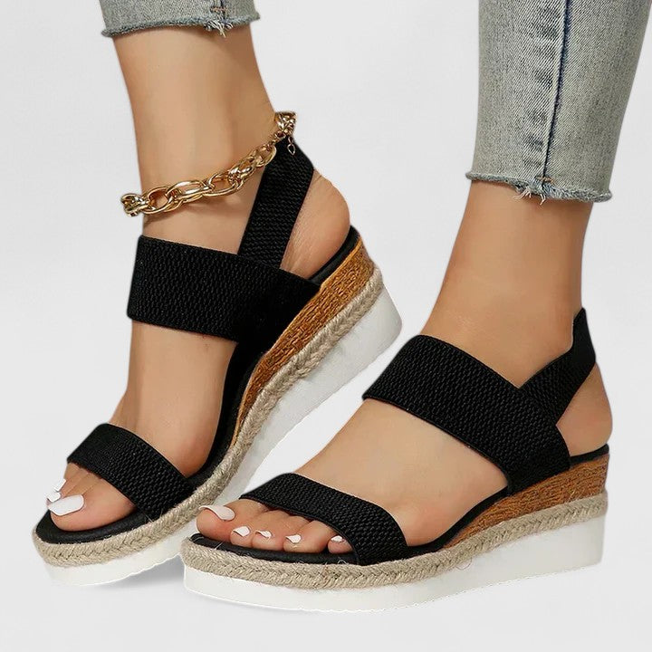 Black wedge sandals with wooden soles worn by a person on a light gray background