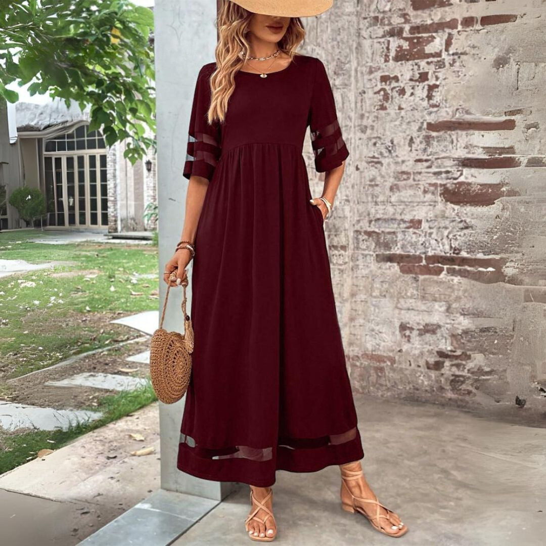 Woman wearing a burgundy dress with sheer sleeves against a brick wall.