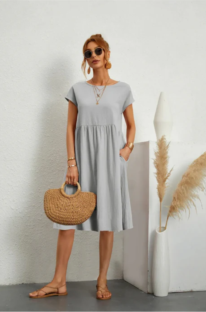 Woman wearing a light gray dress holding a woven bag against a white wall.