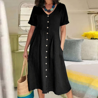 Woman wearing a black dress with buttons, holding a straw bag in a room with a bed and pillows.