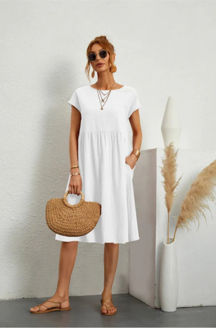 Woman in a white dress holding a straw bag against a plain background