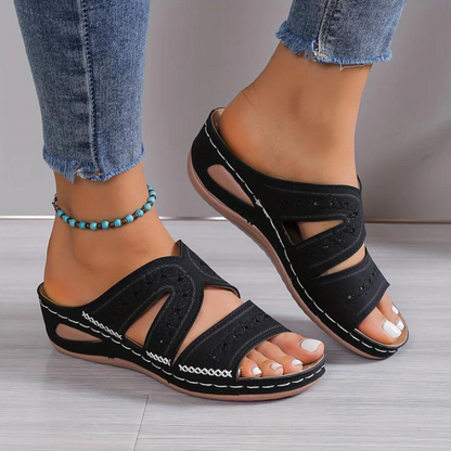 Black sandals worn with blue jeans on a light wooden floor.