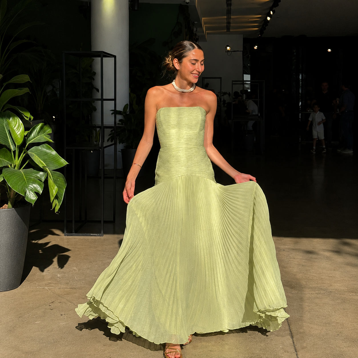 Woman in a light green strapless gown standing outdoors with plants and people in the background.