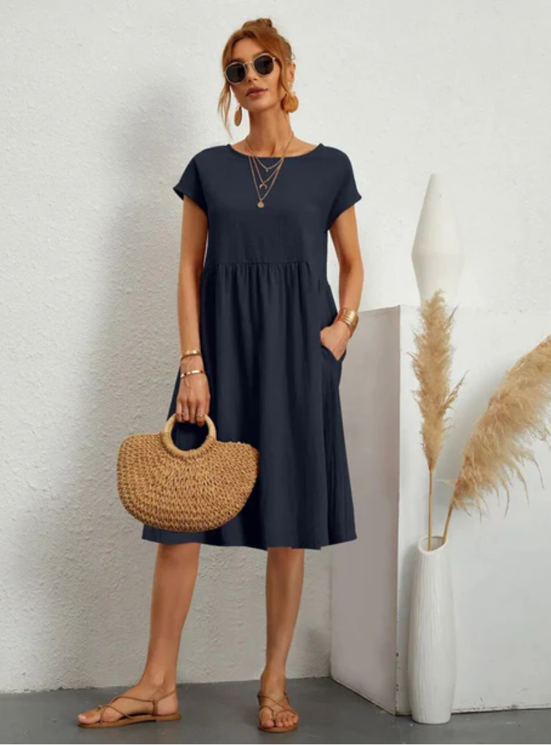 Woman wearing a navy blue dress holding a woven bag against a white wall.
