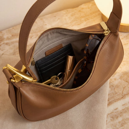 Brown leather shoulder bag with gold zipper partially open, showing keys and a wallet on a beige surface.