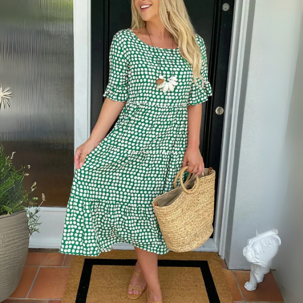 Woman in a green checkered dress holding a straw bag, standing in front of a door.