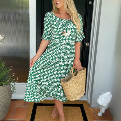 Woman in a green checkered dress holding a straw bag, standing in front of a door.