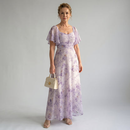 Woman wearing a purple floral dress holding a beige handbag against a plain background