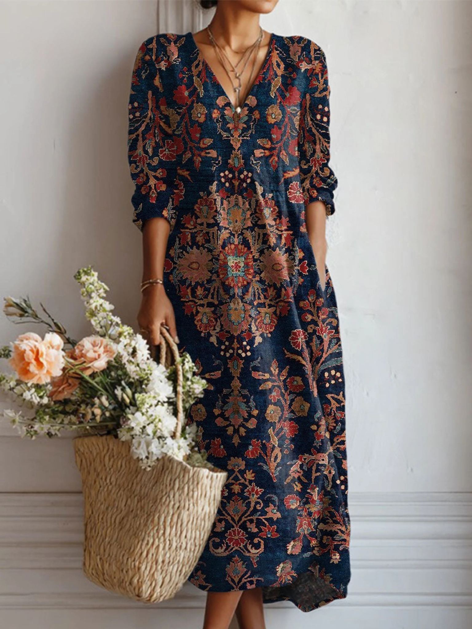 Woman wearing a long, patterned dress holding a woven bag with flowers against a white wall.