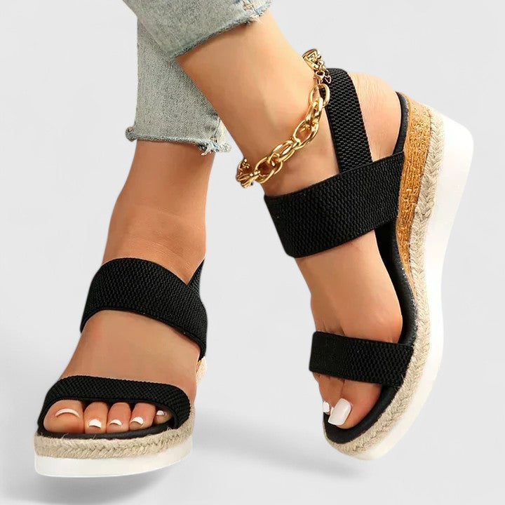 Black wedge sandals with cork sole worn by a person on a light gray background