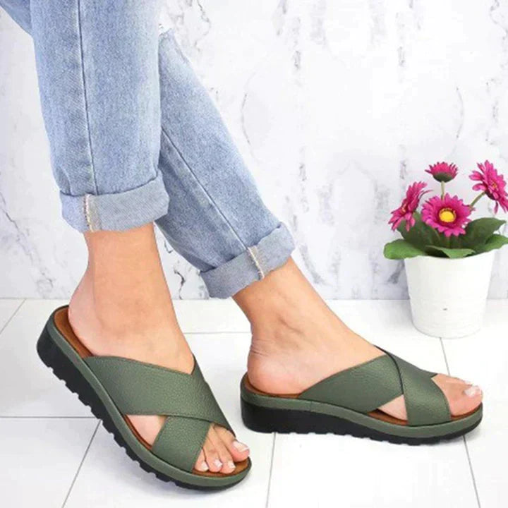Green sandals worn with light blue jeans on a white floor with a plant in the background