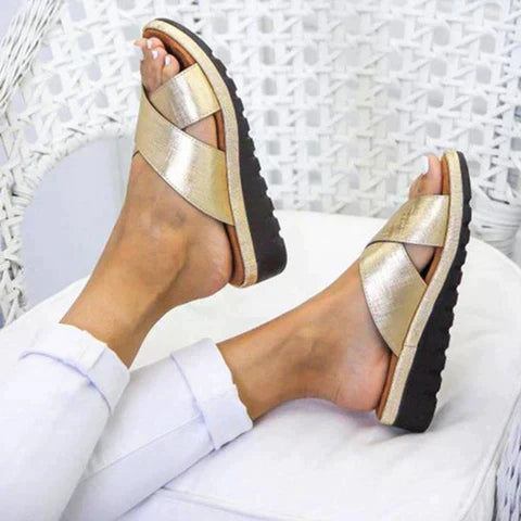 Gold sandals with black soles worn by a person sitting on a white chair.
