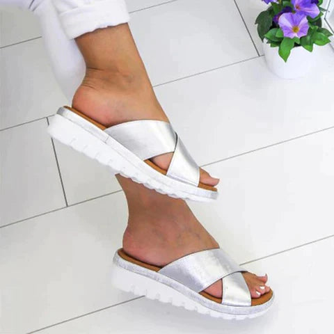 White sandals worn on a white tiled floor with a plant in the corner.