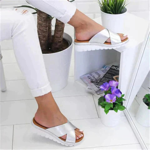 White sandals worn by a person on a white floor with plants and a magazine.
