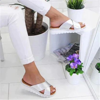 White sandals worn by a person on a white floor with plants and a magazine.