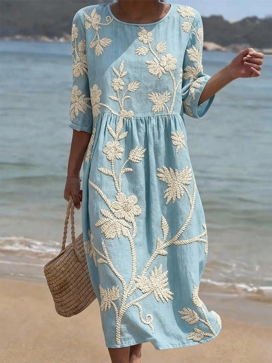 Woman wearing a light blue dress with white floral embroidery by the beach.