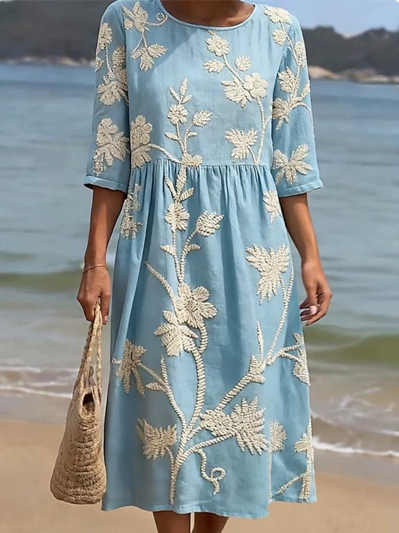 Woman wearing a light blue dress with white floral embroidery on a beach.