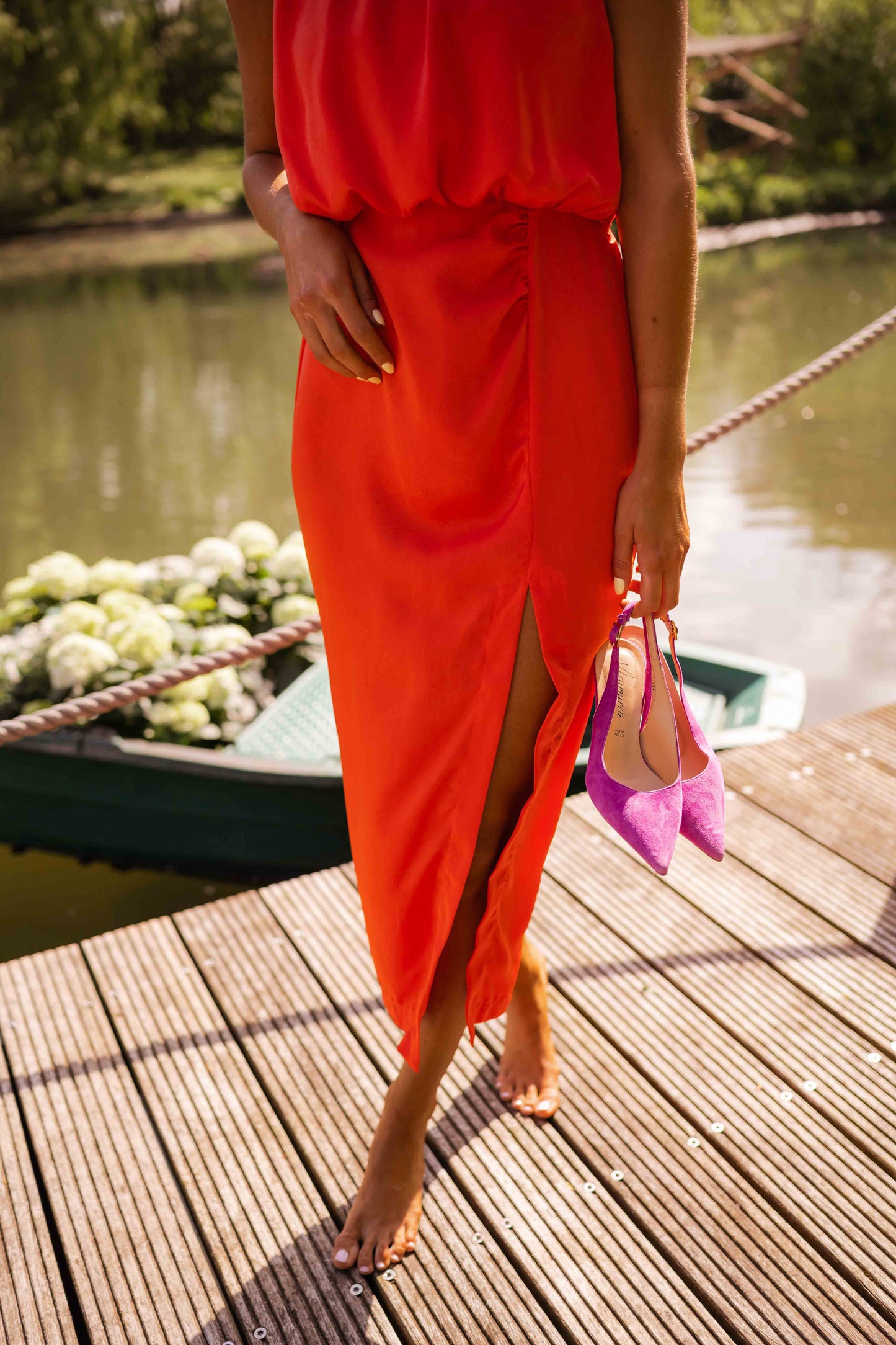 Person wearing a bright orange dress with a slit, standing on a wooden dock by a body of water.